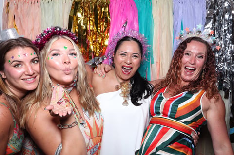 Four smiling women at a party with colorful decorations and sequined backdrops, wearing festive attire with face glitter and crowns