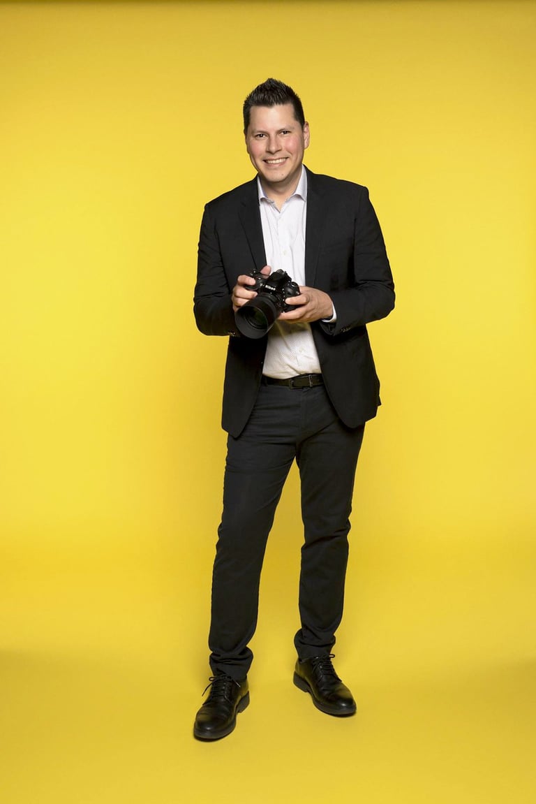 Man in black suit and white shirt holding a camera against a bright yellow background