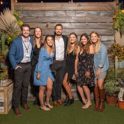 Seven people posing together in front of a wooden wall with decorative plants
